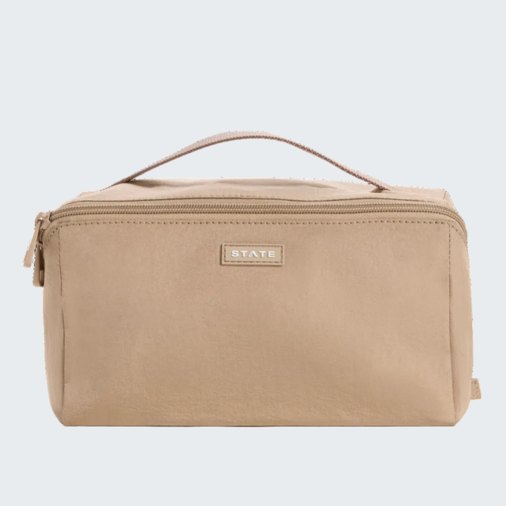 A beige canvas toiletry bag with a top handle and a small metallic brand plaque reading 'STAYE' on the front, shown against a plain white background.