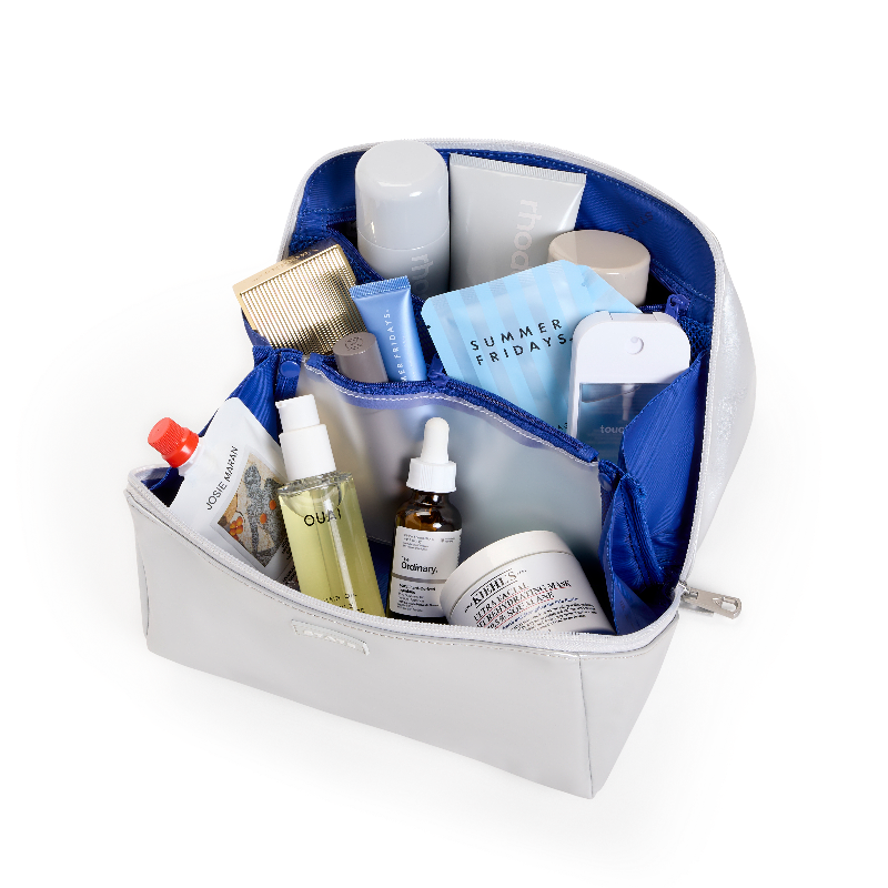 A light gray zippered cosmetic bag shown open on a white background, filled with assorted skincare and beauty products including bottles, tubes, a comb, and a phone case.