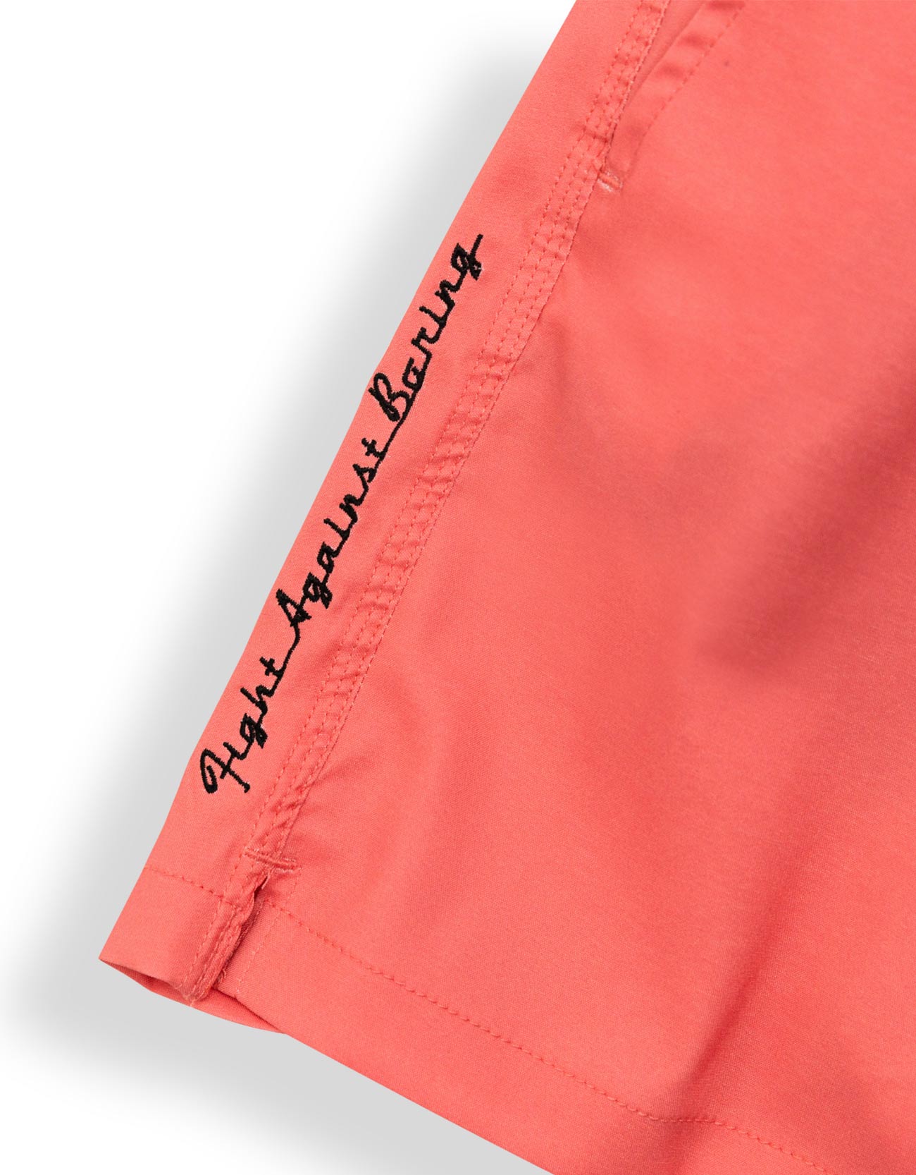Close-up of a coral pink garment sleeve with black script embroidery reading 'Fight Against Boring' on the cuff, shown against a white background.