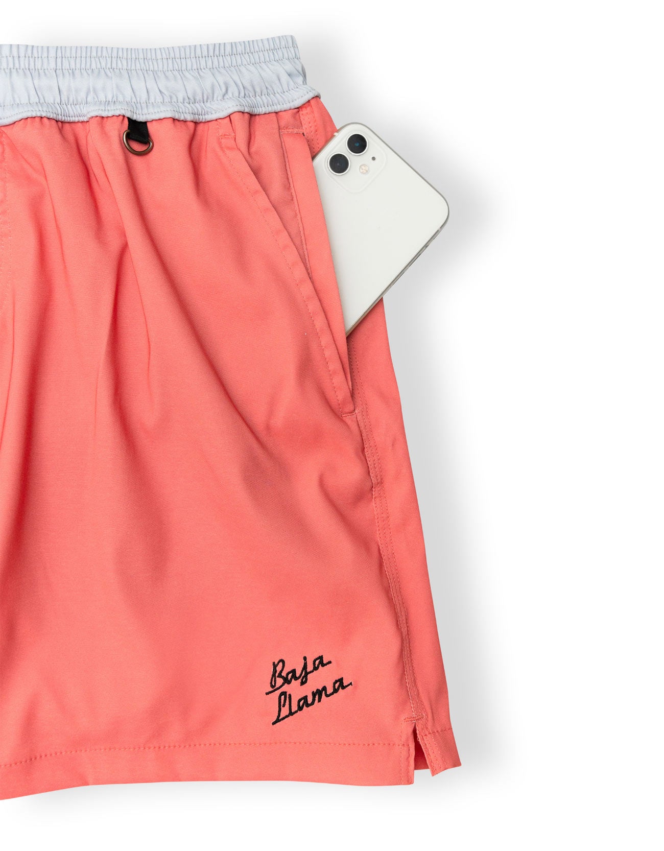 Coral pink drawstring shorts with a light gray elastic waistband and black 'Baja Lama' script embroidery near the hem, shown with a white smartphone tucked into the side pocket on a white background.