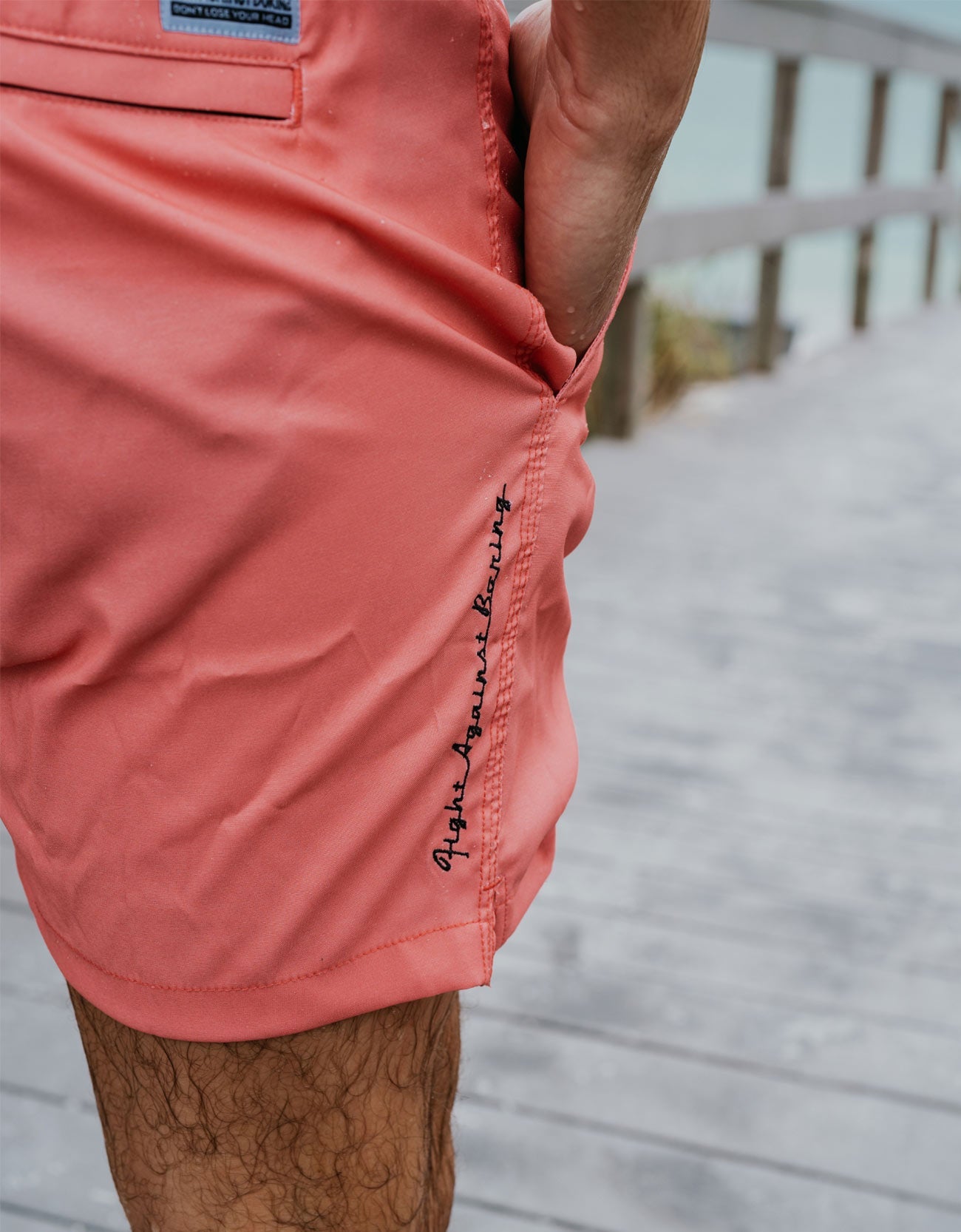 Close-up of a person wearing coral pink board shorts with black script embroidery on the side, standing on a wooden boardwalk.