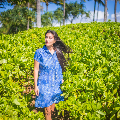 Woman in a blue dress standing in a lush green field with palm trees in the background