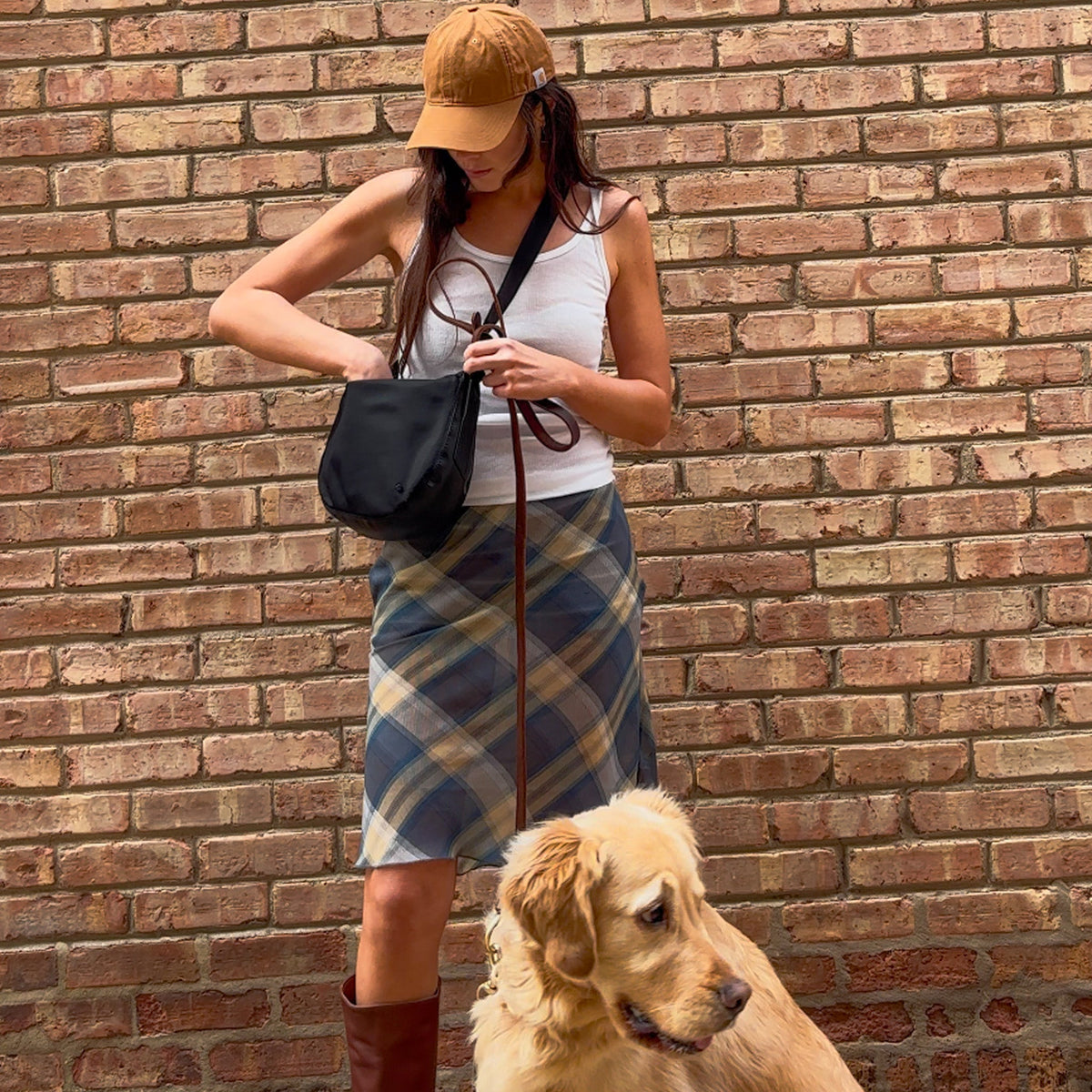 Woman in plaid skirt and tan cap with a dog against a brick wall