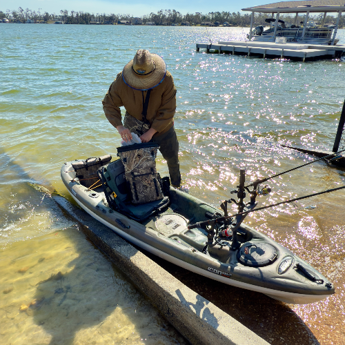 Person preparing a kayak on a lakeside with fishing equipment.