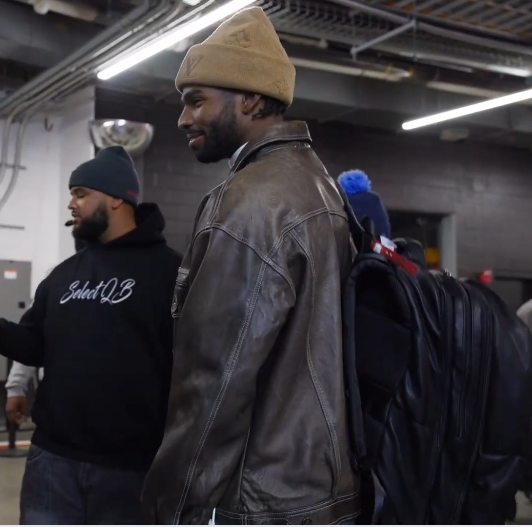 Two men walking in an indoor setting, one wearing a leather jacket and beanie, the other in a black hoodie with text.