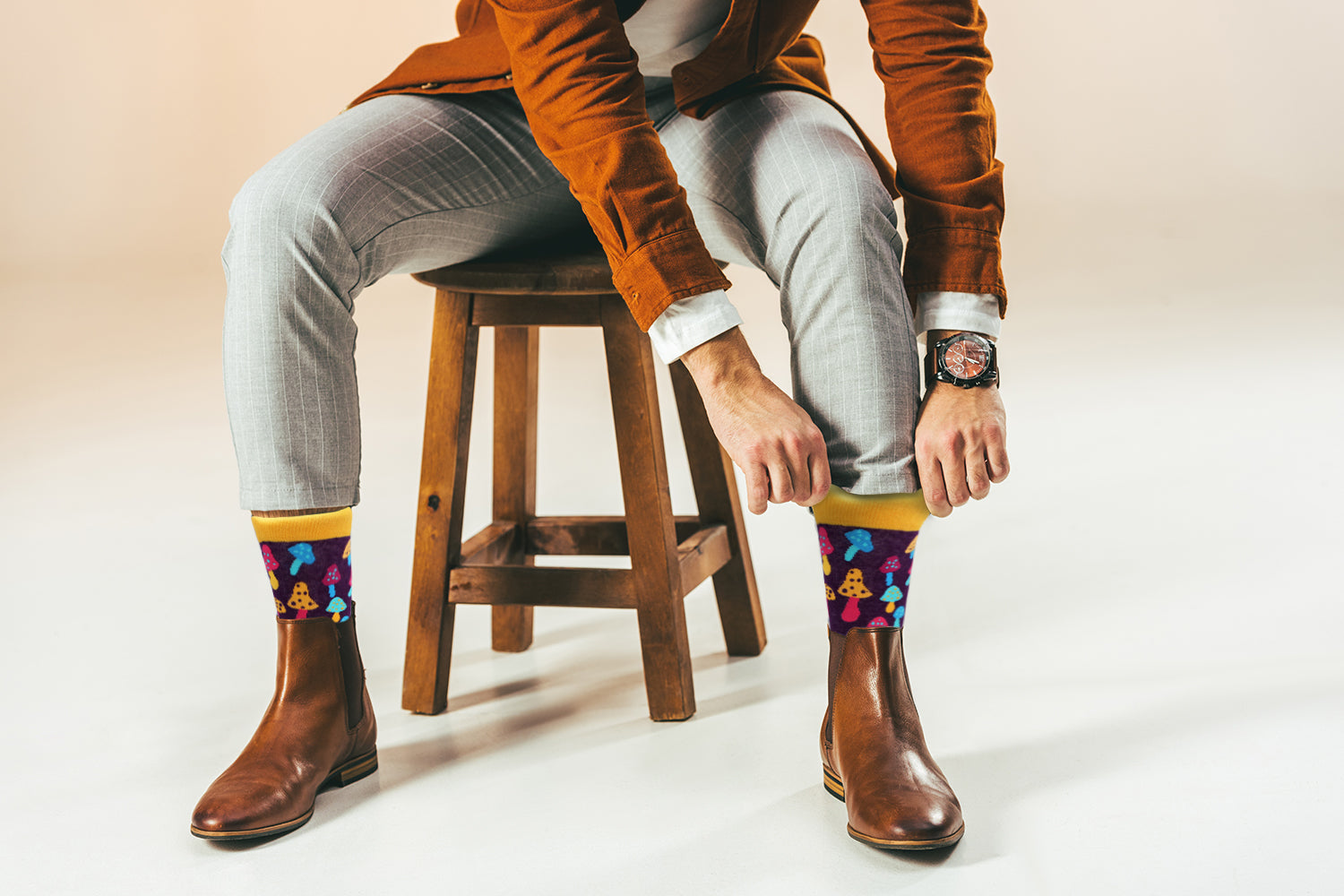 Person sitting on a wooden stool wearing colorful socks and a brown jacket.