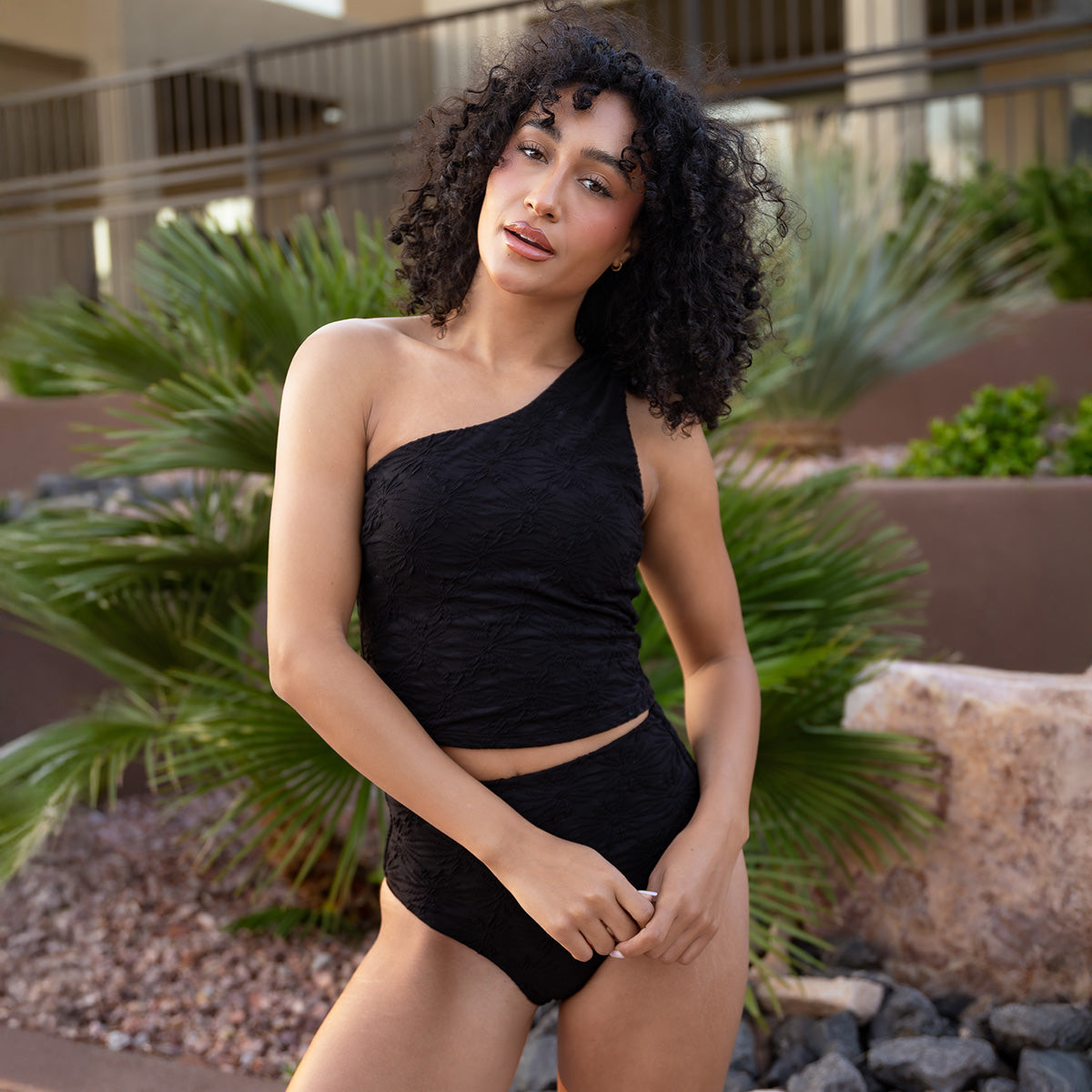 Woman in a black one-shoulder top and shorts standing outdoors with plants in the background