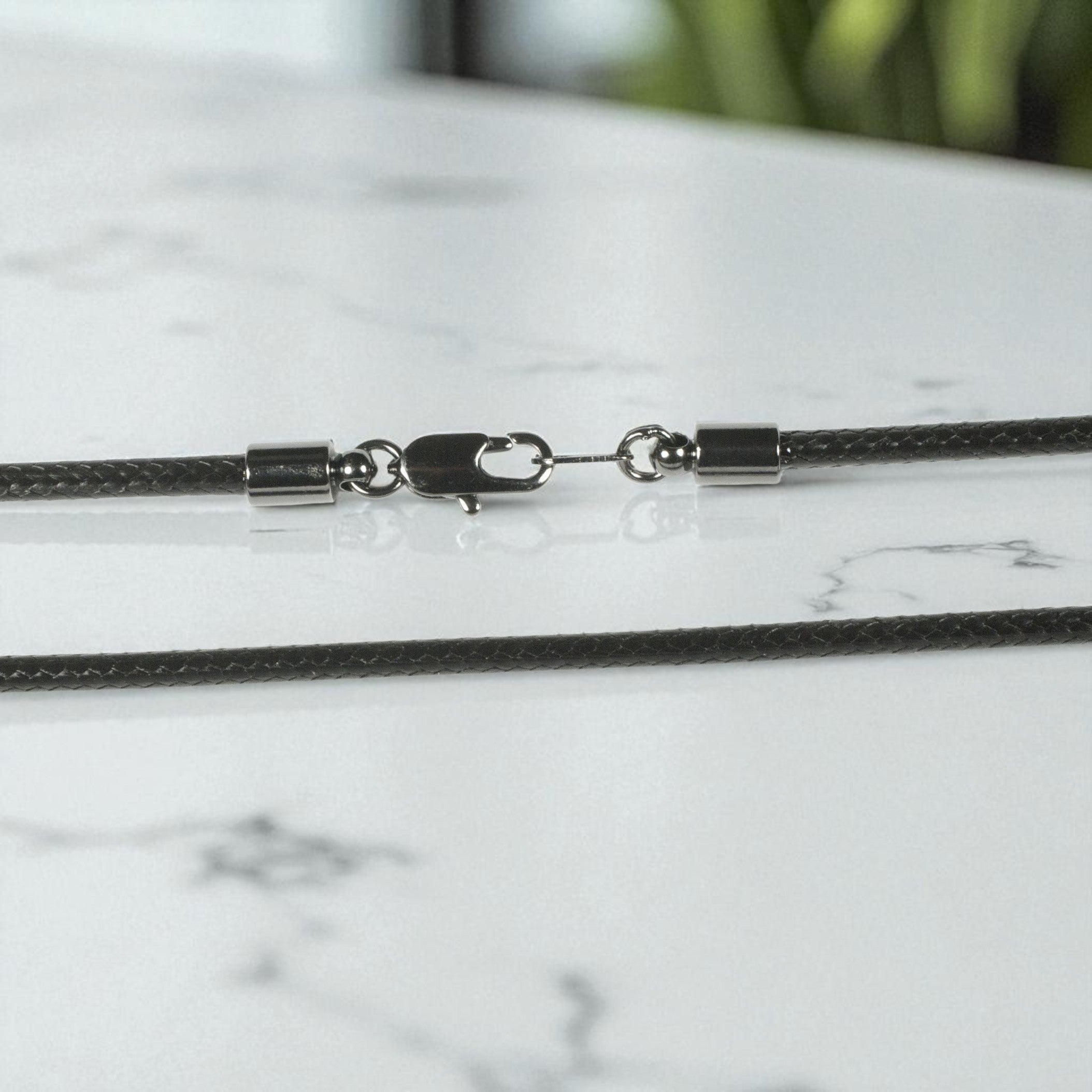 A close-up of a dark gray braided chain necklace with a silver-toned lobster clasp and metal end caps, resting on a white marble surface.