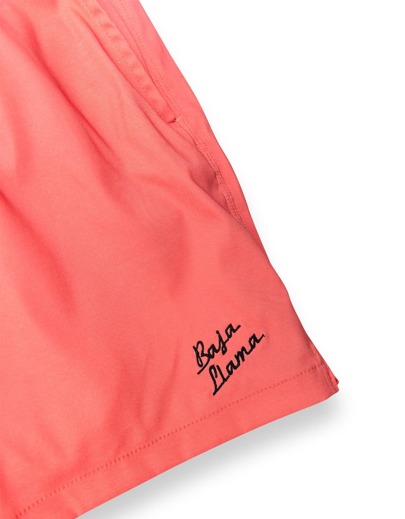 Close-up of a coral pink garment, likely shorts or a skirt, with black script embroidery reading 'Baba Yana' near the hem on a white background.