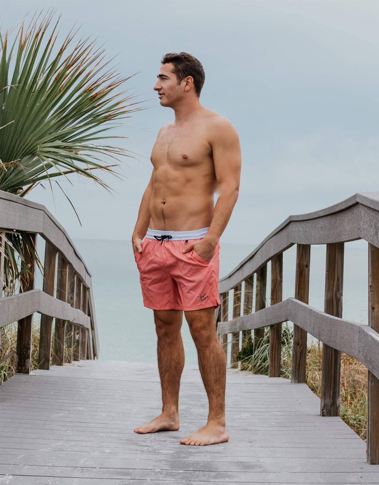 A man stands on a wooden boardwalk by the beach wearing coral pink swim trunks with a white waistband. Palm leaves and a cloudy sky are visible in the background.