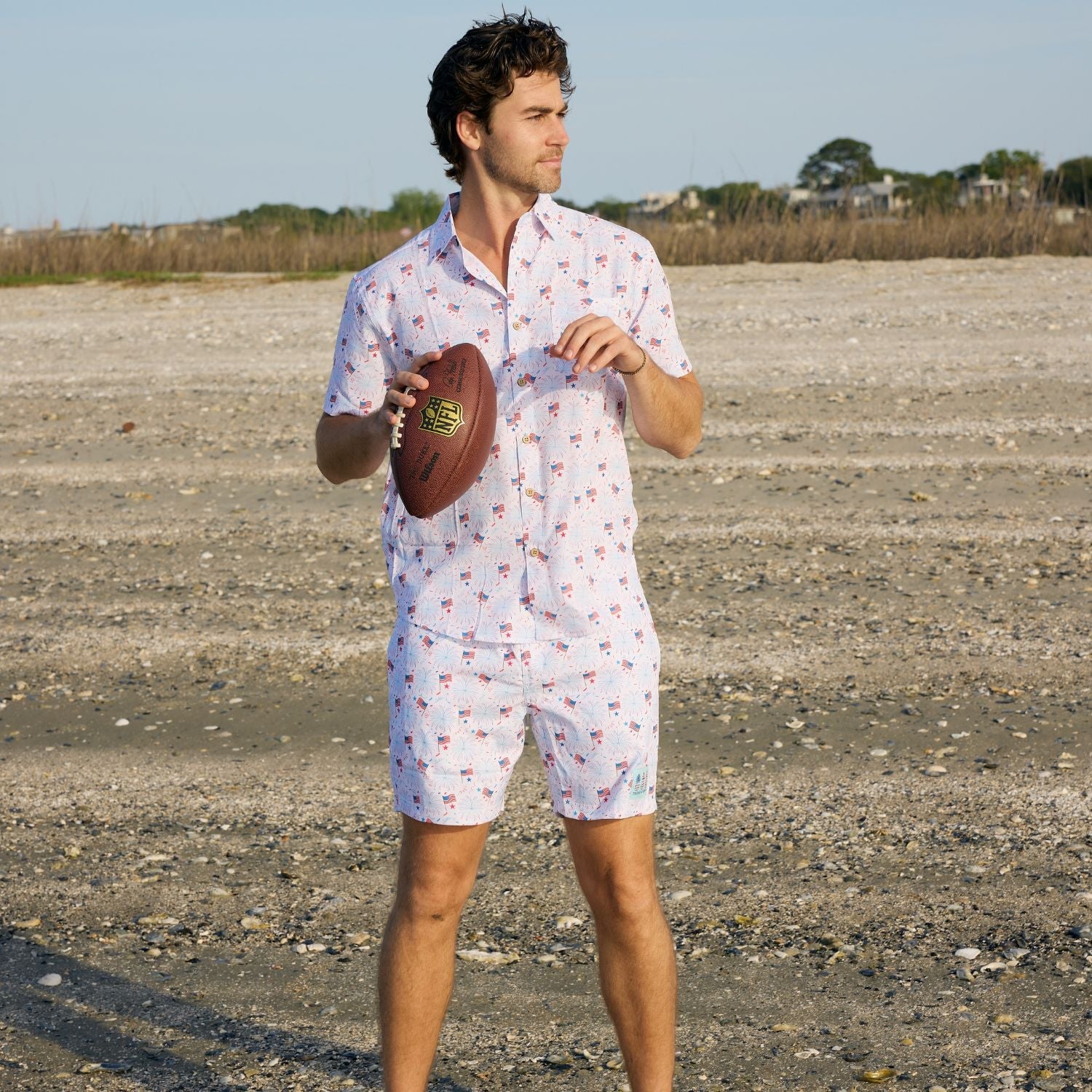 Man in a patterned shirt and shorts holding a football on a gravel road.