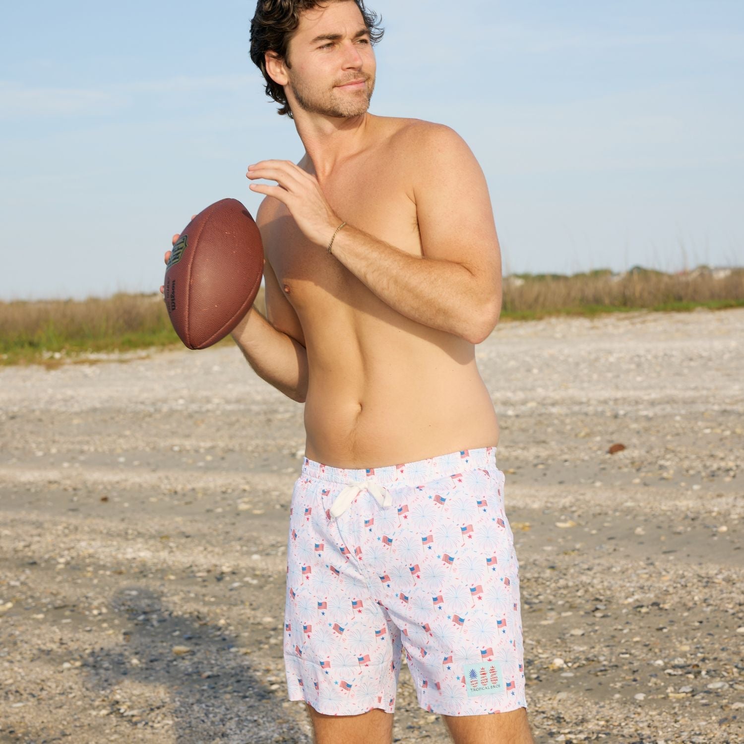 Man holding a football on a beach wearing patterned shorts