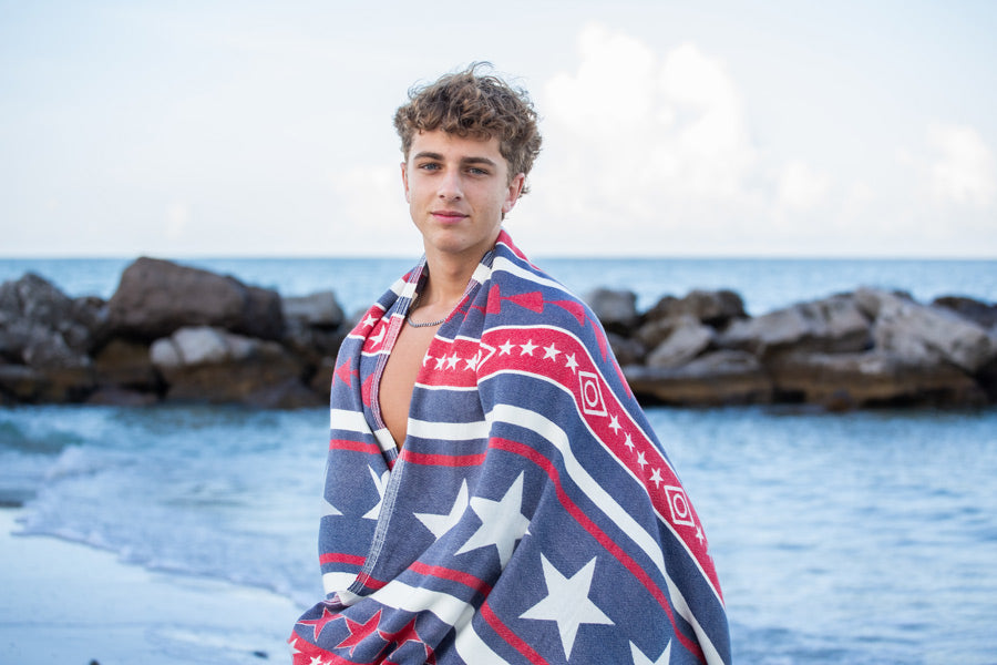 Person wrapped in a red, white, and blue patterned towel on a beach with rocks and water in the background