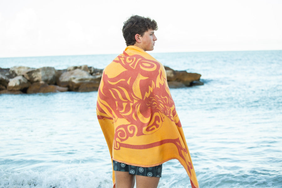 Person wrapped in a yellow towel with red patterns standing by the ocean.