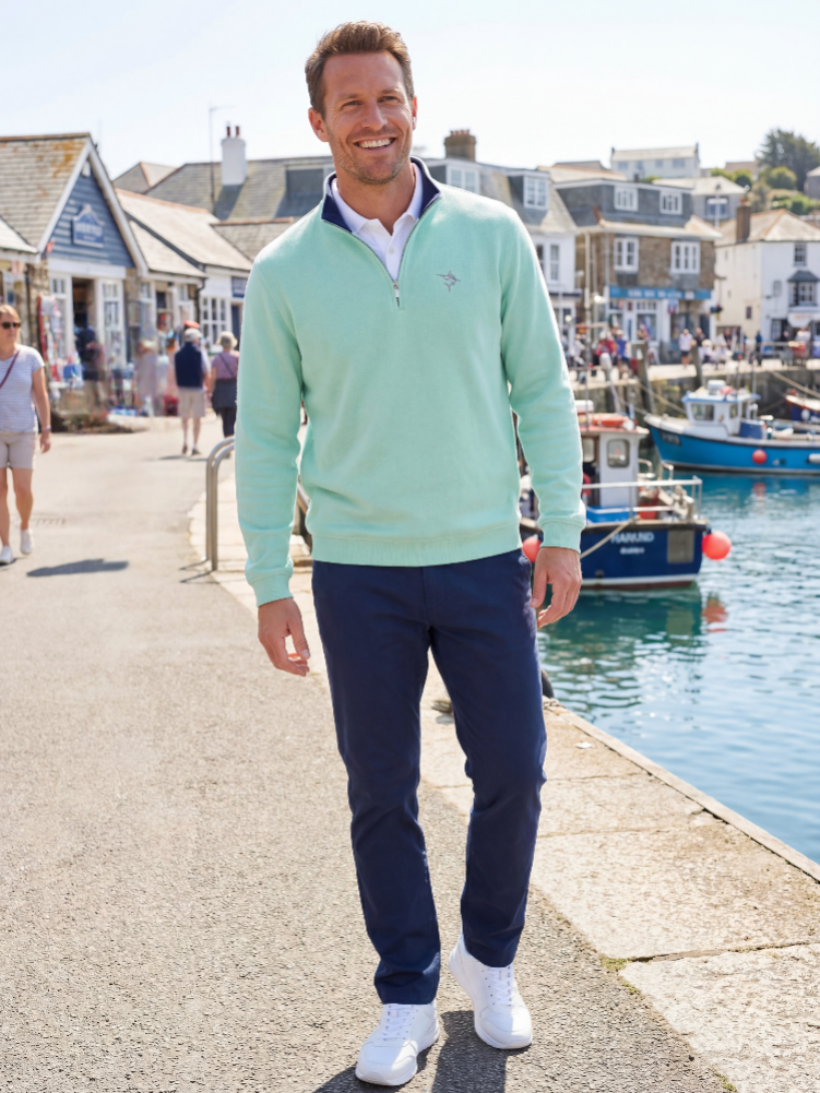 Man in a light green sweater and navy pants standing by a harbor with buildings and boats in the background.