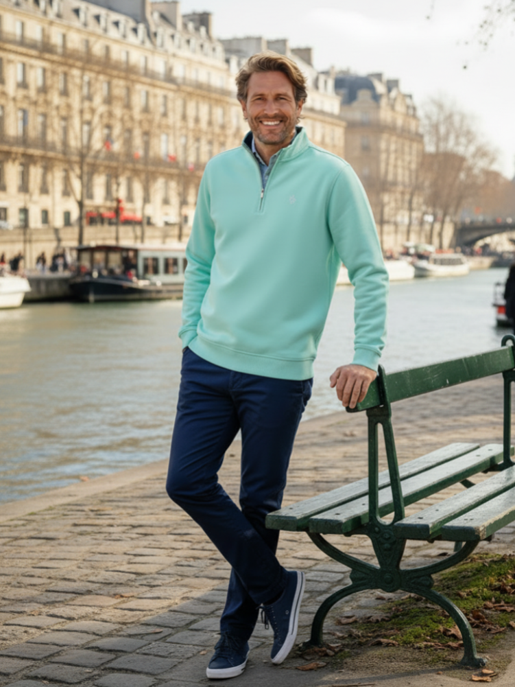 Man in a light blue sweater and dark pants standing by a river with a bench in the foreground.