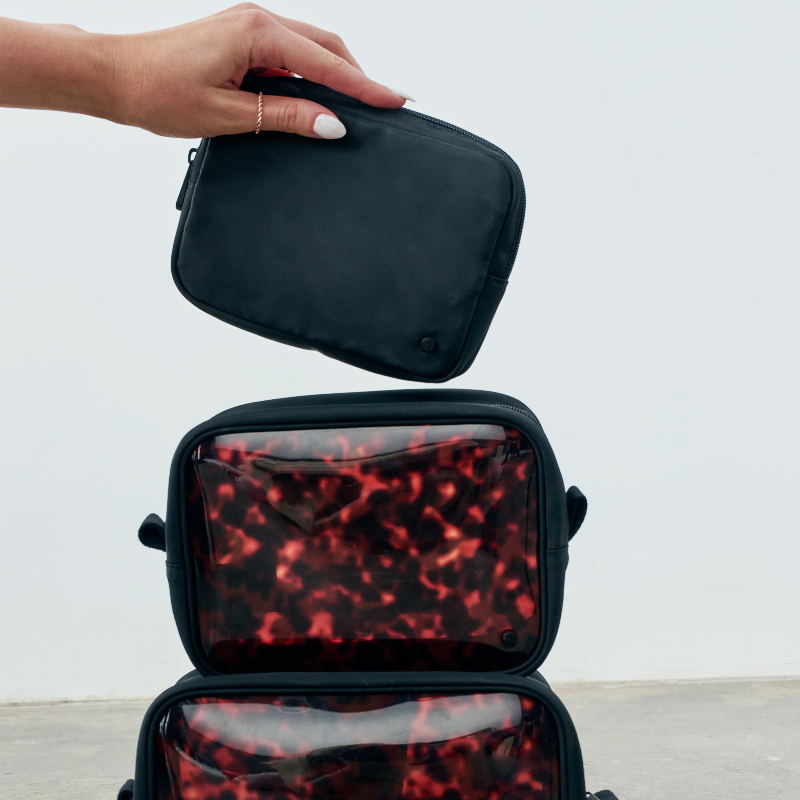 A hand holds a black zip pouch above two stacked translucent toiletry bags with a red and black marbled pattern, against a plain light background.