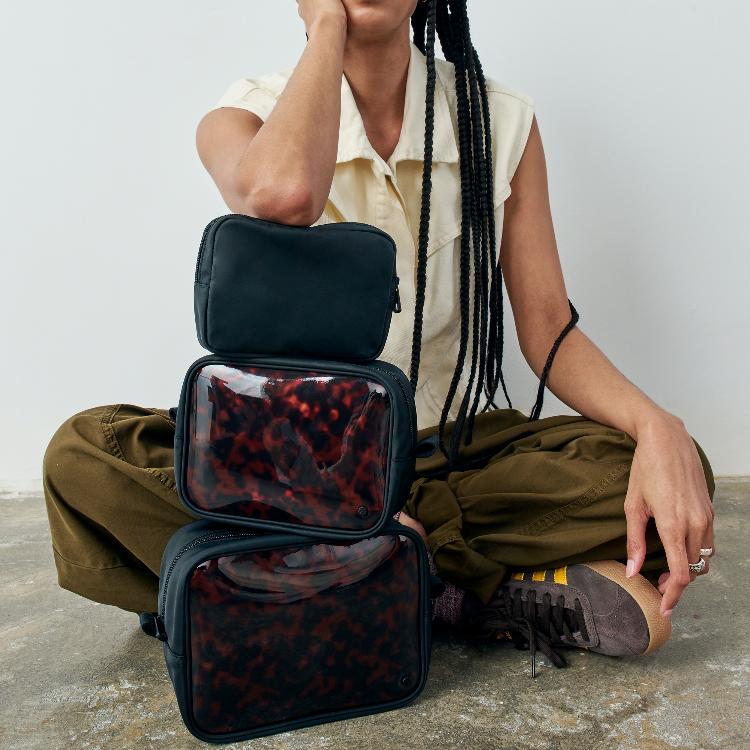 A person sits cross-legged on a concrete floor against a light gray wall, wearing olive green pants and a cream sleeveless top, with three stacked black travel organizer pouches in front. The middle and bottom pouches have clear windows showing a red-and-black tortoiseshell pattern inside.