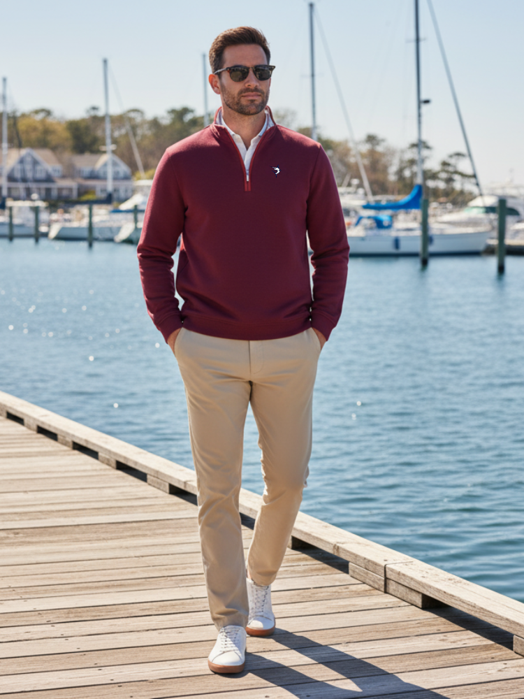 Man in a red sweater and beige pants standing on a dock by a body of water.