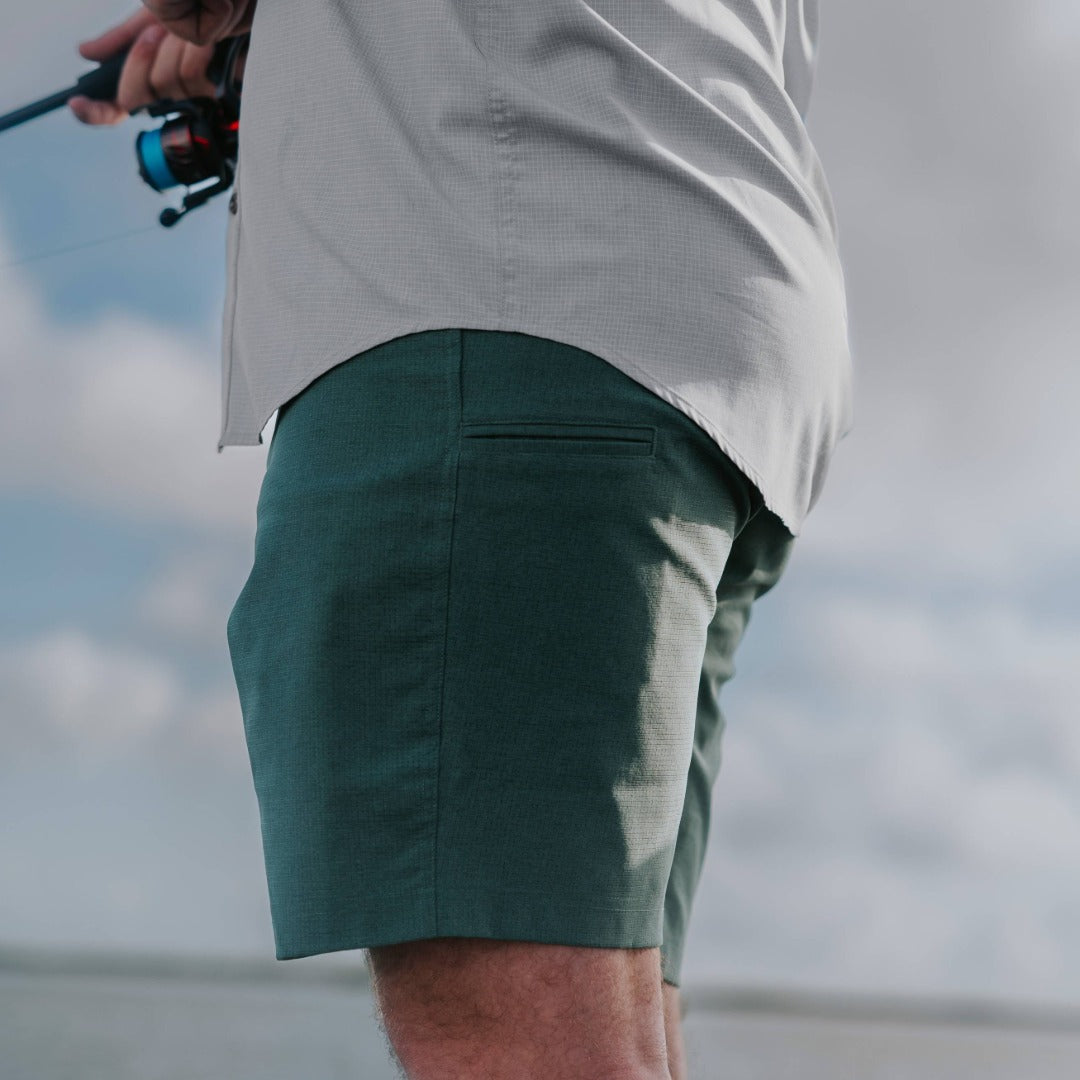 A close-up side view of a person outdoors wearing dark teal-green shorts and a light gray button-up shirt, holding a fishing rod with a blue and black reel against a cloudy sky background.