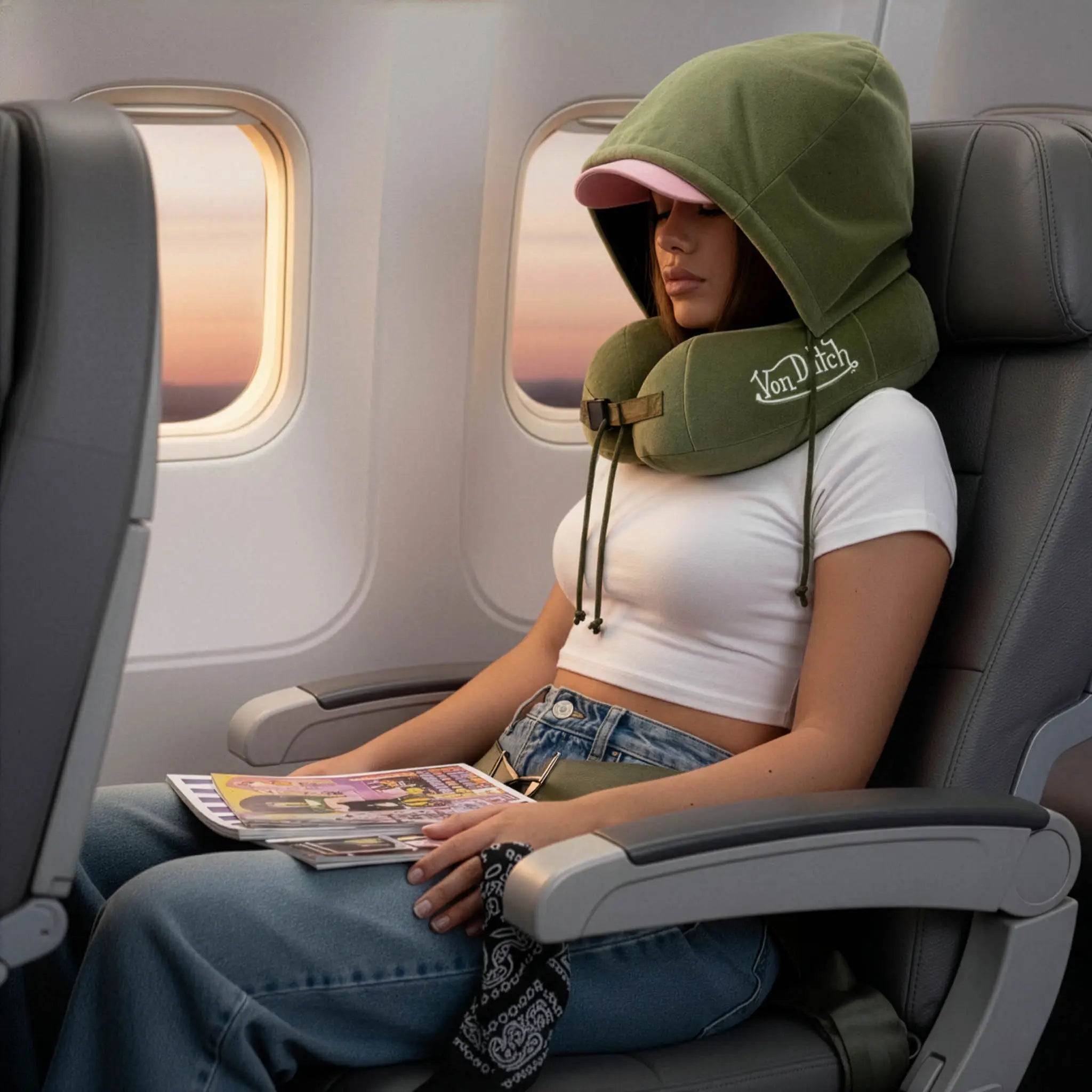 Person sitting in an airplane seat with a green neck pillow, holding a magazine.