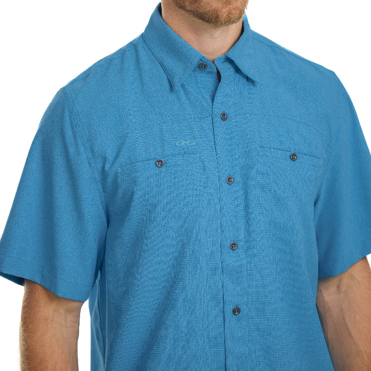 A man wearing a short-sleeve button-up shirt in a heathered blue color, featuring two chest pockets and a small embroidered logo on the left chest.
