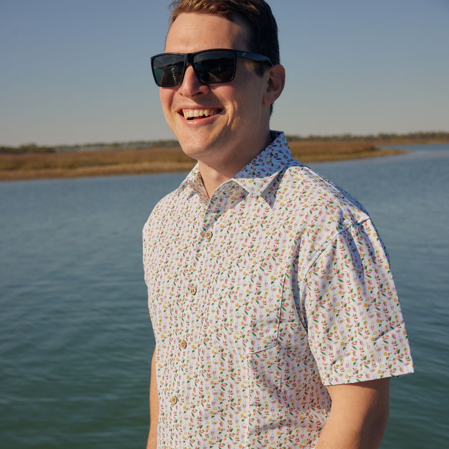 Man wearing sunglasses and a patterned shirt standing by a body of water.