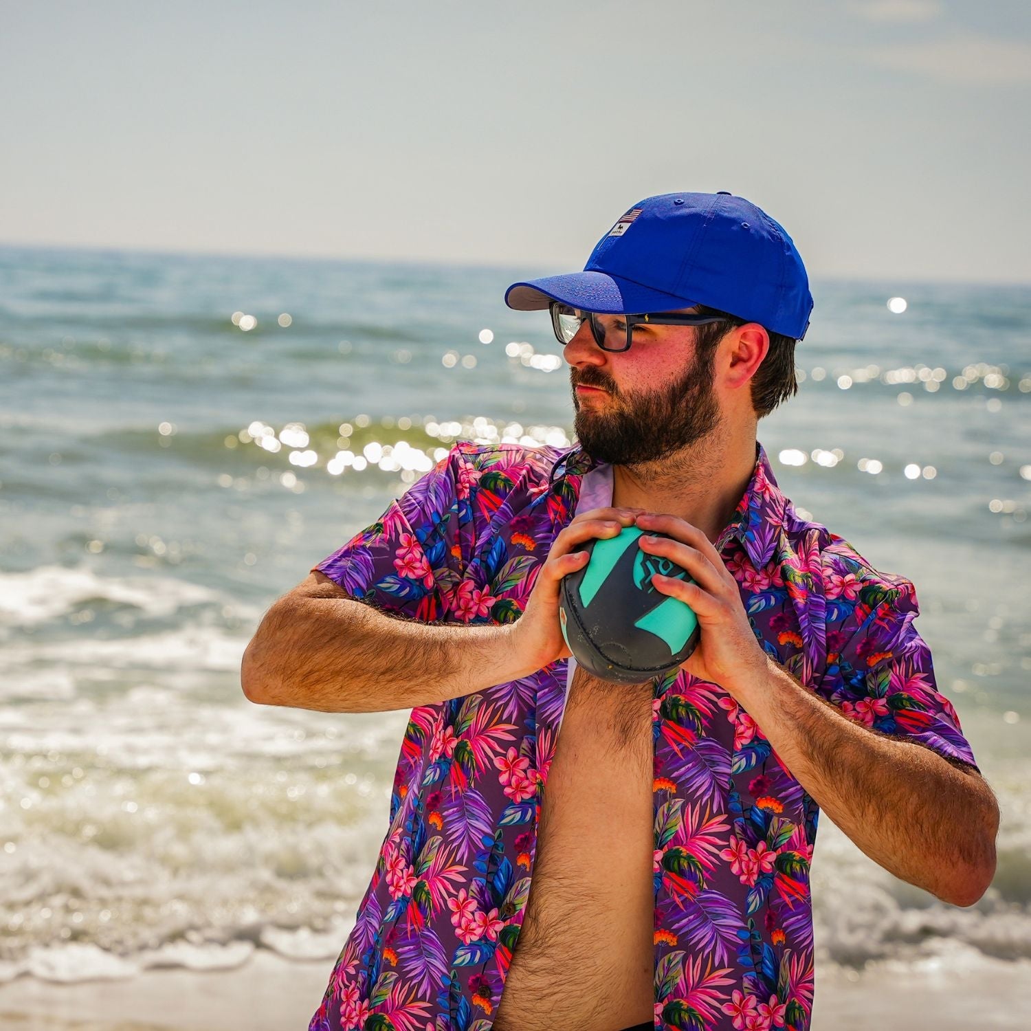 Man in a colorful shirt and blue cap holding a green ball on a beach.