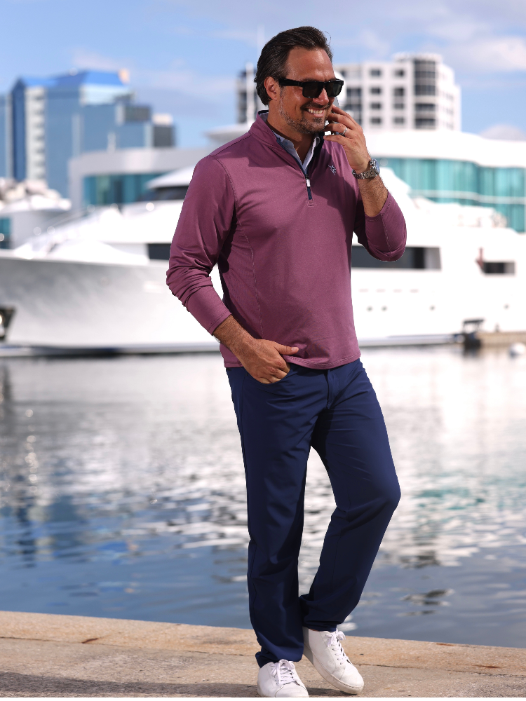 Man in a purple long-sleeve shirt and blue pants standing by a waterfront with boats and buildings in the background.