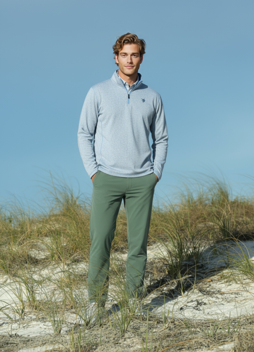 Man wearing a light blue pullover and green pants standing on sandy dunes with a clear blue sky.