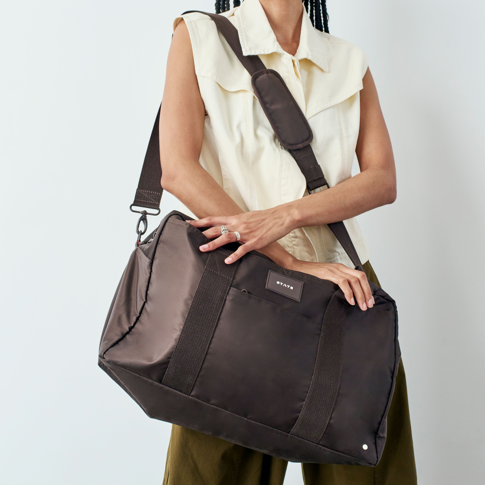 Person holding a brown duffel bag with a visible brand logo on a light gray background