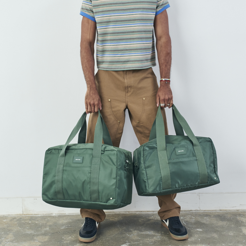 Person holding two green duffel bags against a plain background