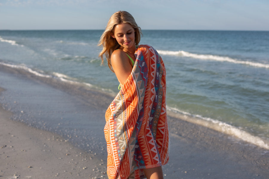 Woman on a beach wrapped in an orange patterned towel with ocean waves in the background
