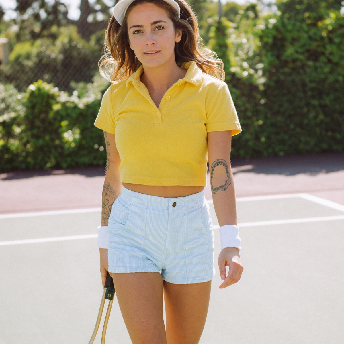 Woman in yellow polo shirt and light blue shorts holding a tennis racket on a tennis court.