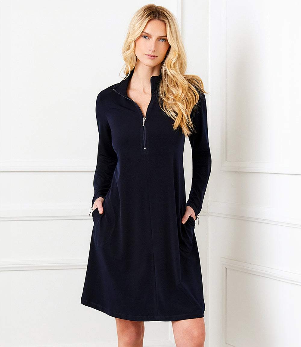A blonde woman modeling a dark navy blue long-sleeve shift dress with a half-zip mock neck and side pockets, standing against a white paneled wall.