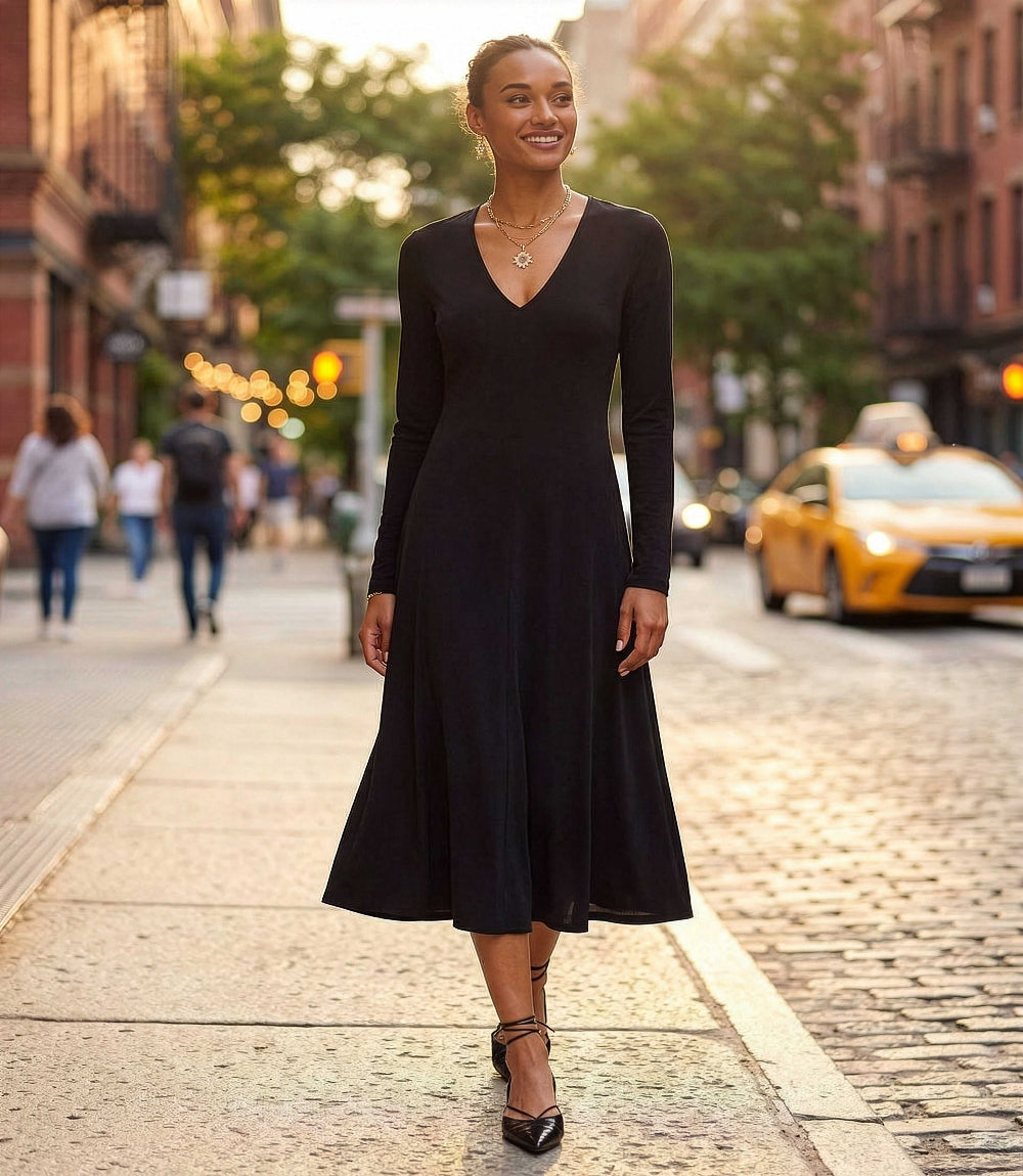 A woman wearing a black long-sleeve midi dress with a V-neckline walks down a cobblestone city street at sunset. A yellow taxi is visible in the background, making this a lifestyle shot.