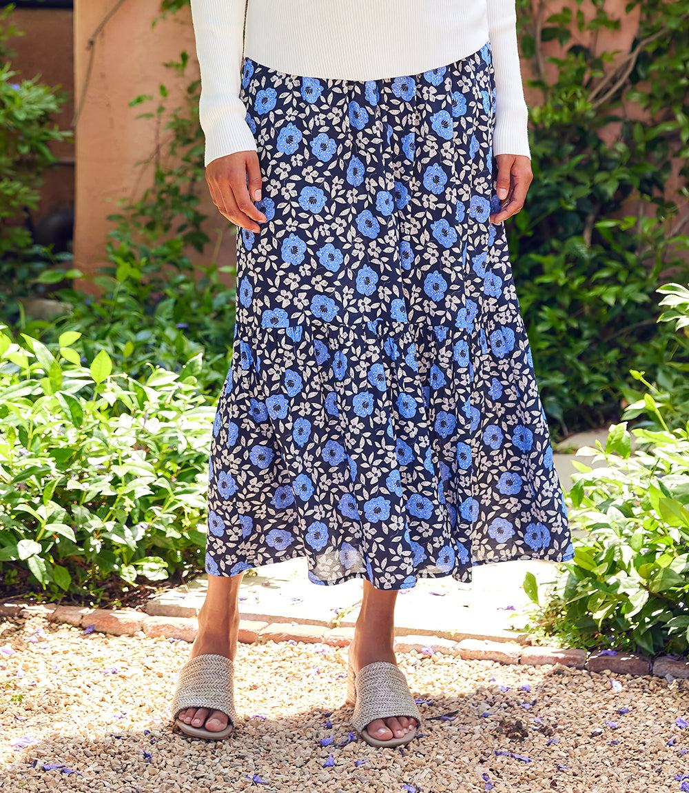 A person wearing a navy midi skirt with a blue and white floral print, paired with a white long-sleeve top and gray slide sandals, standing outdoors in a garden setting.