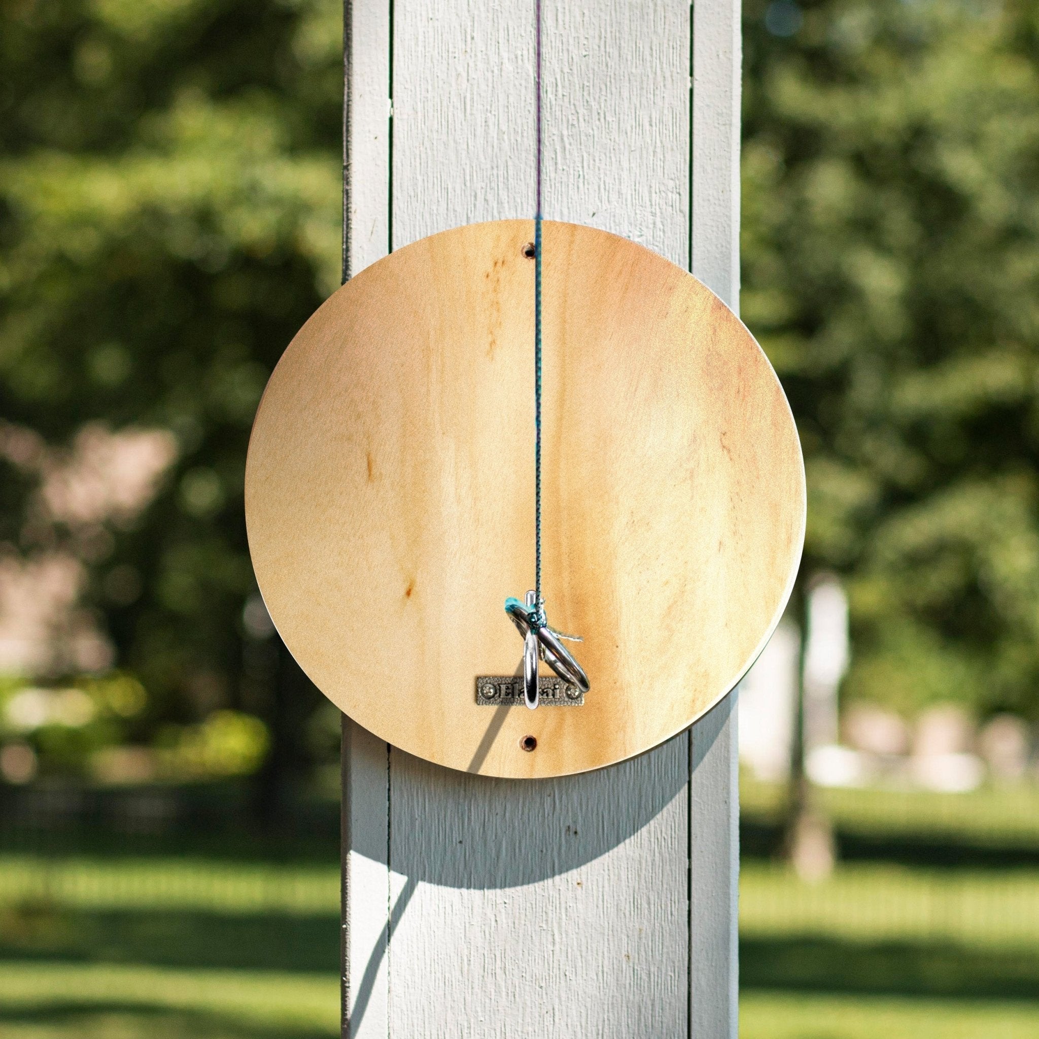 A circular wooden fly tying stand with a hanging string and small metal pegs, mounted on a white wooden post in an outdoor garden setting.