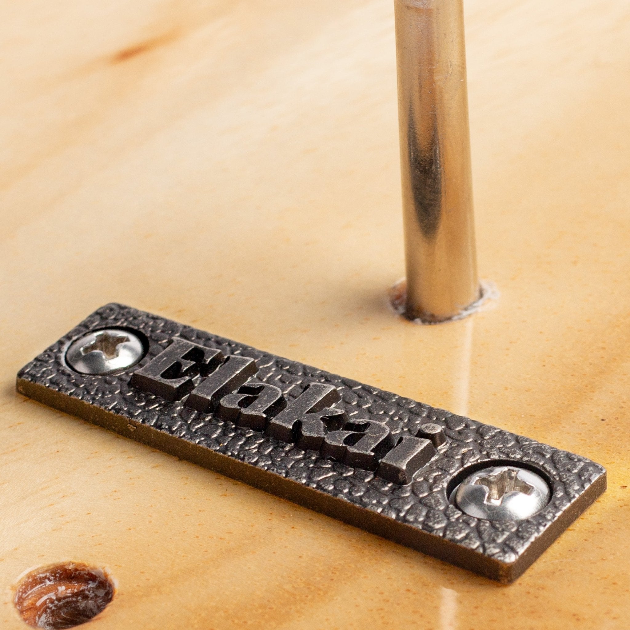 A close-up of a rectangular metal nameplate with the name 'Elakai' raised in the center, mounted on a light wood surface next to a vertical metal post.