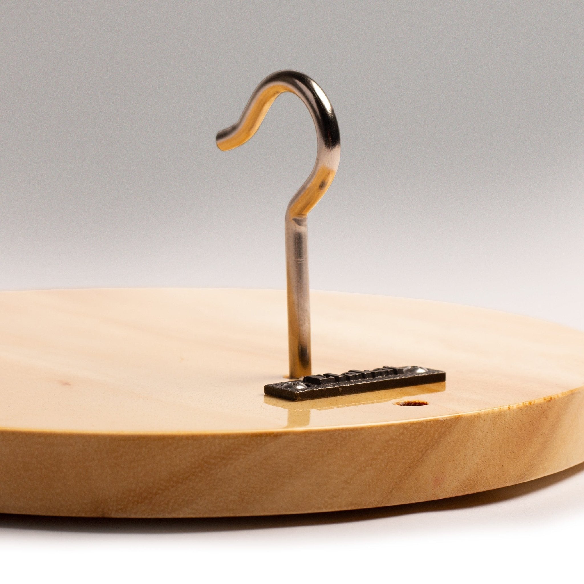 A close-up of a light natural wood round board with a polished metal hook mounted on a small black base, photographed against a soft gray studio background.