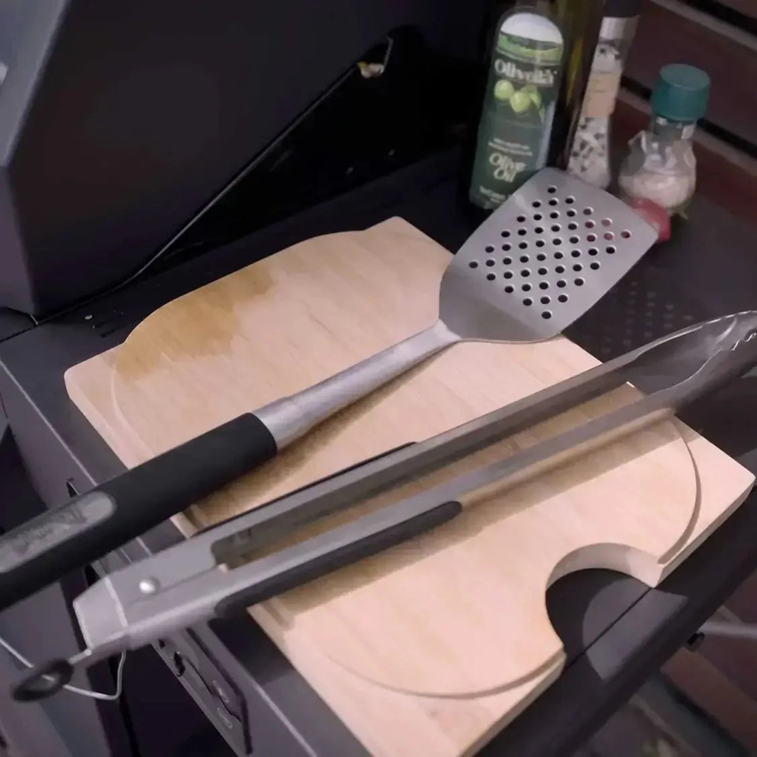 Barbecue tools including tongs and a spatula on a cutting board with a grill in the background.