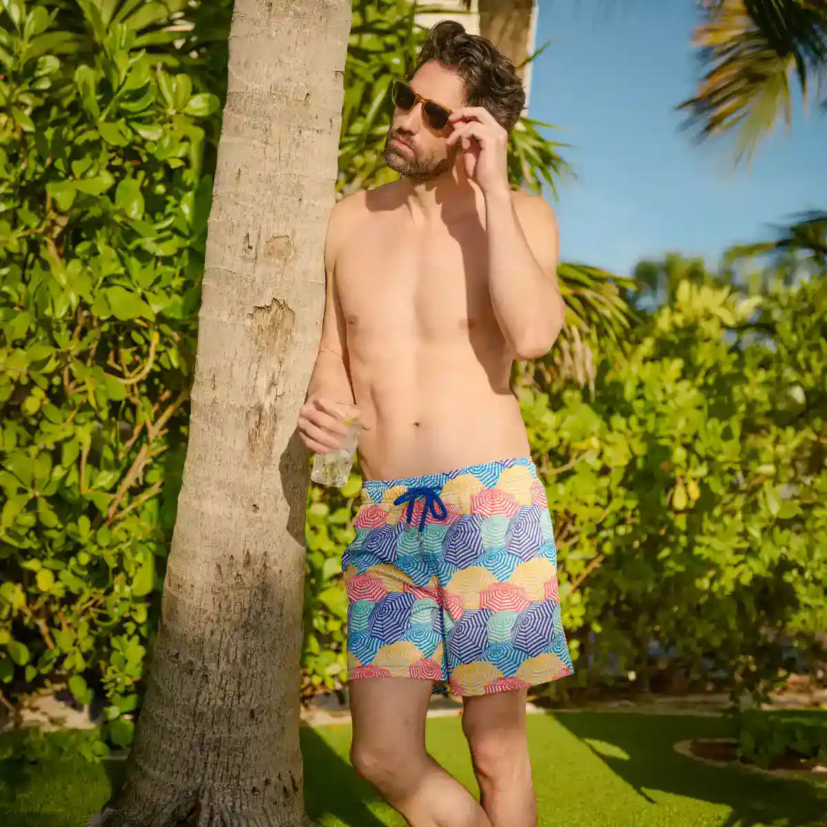 Man wearing colorful swim shorts standing next to a palm tree in a tropical setting