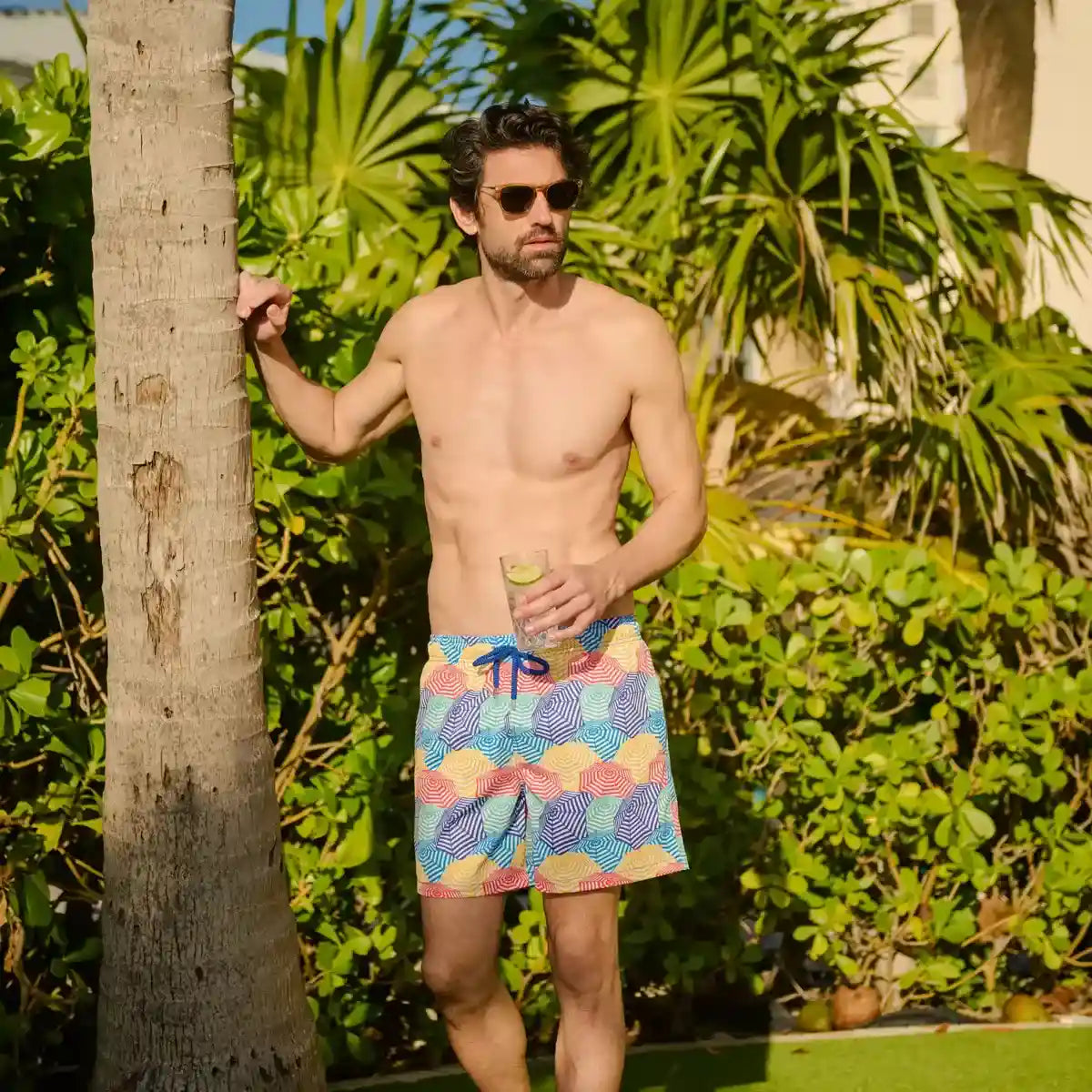 Man wearing colorful swim shorts standing outdoors with greenery in the background