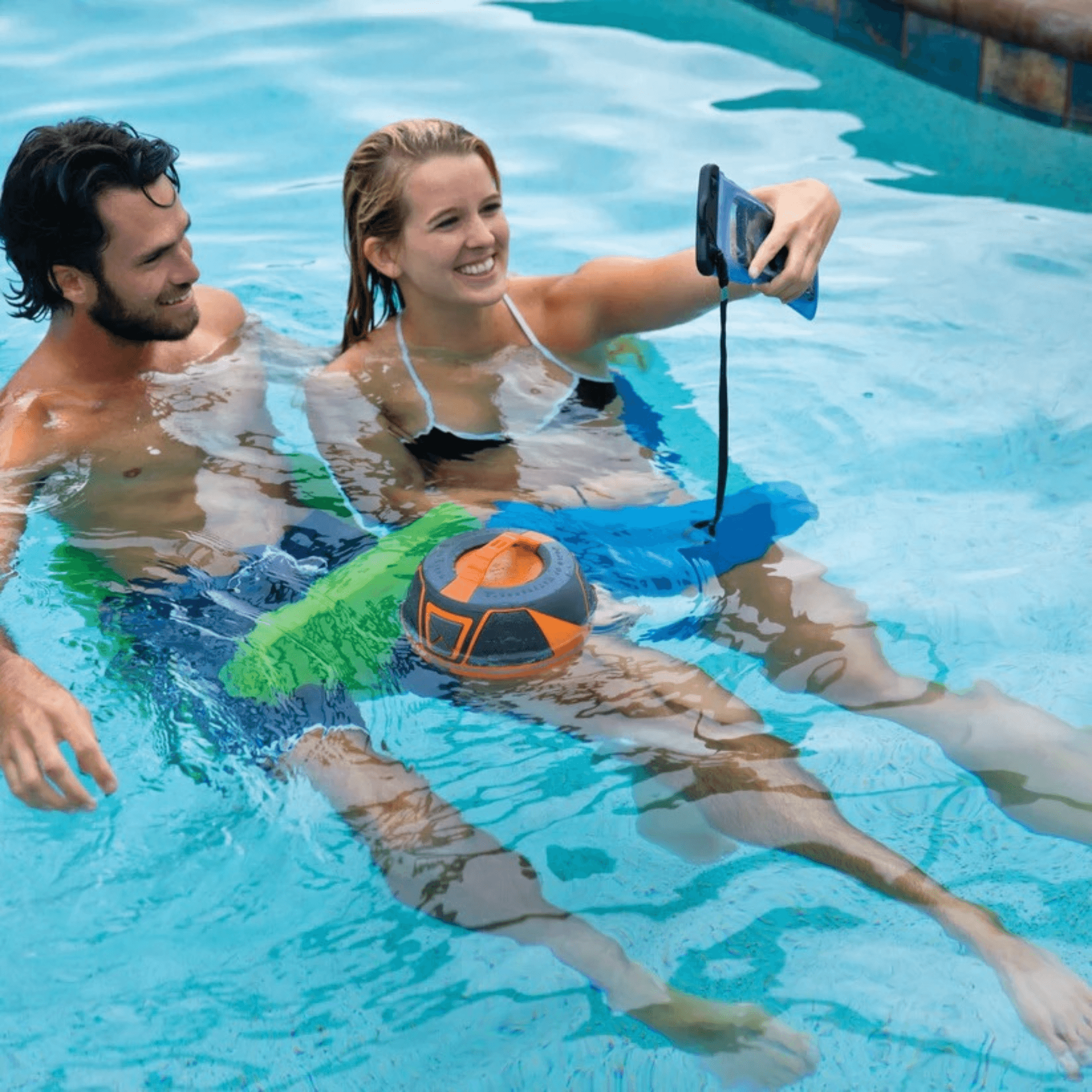 Two people in a pool with a ball and smartphone, taking a selfie.