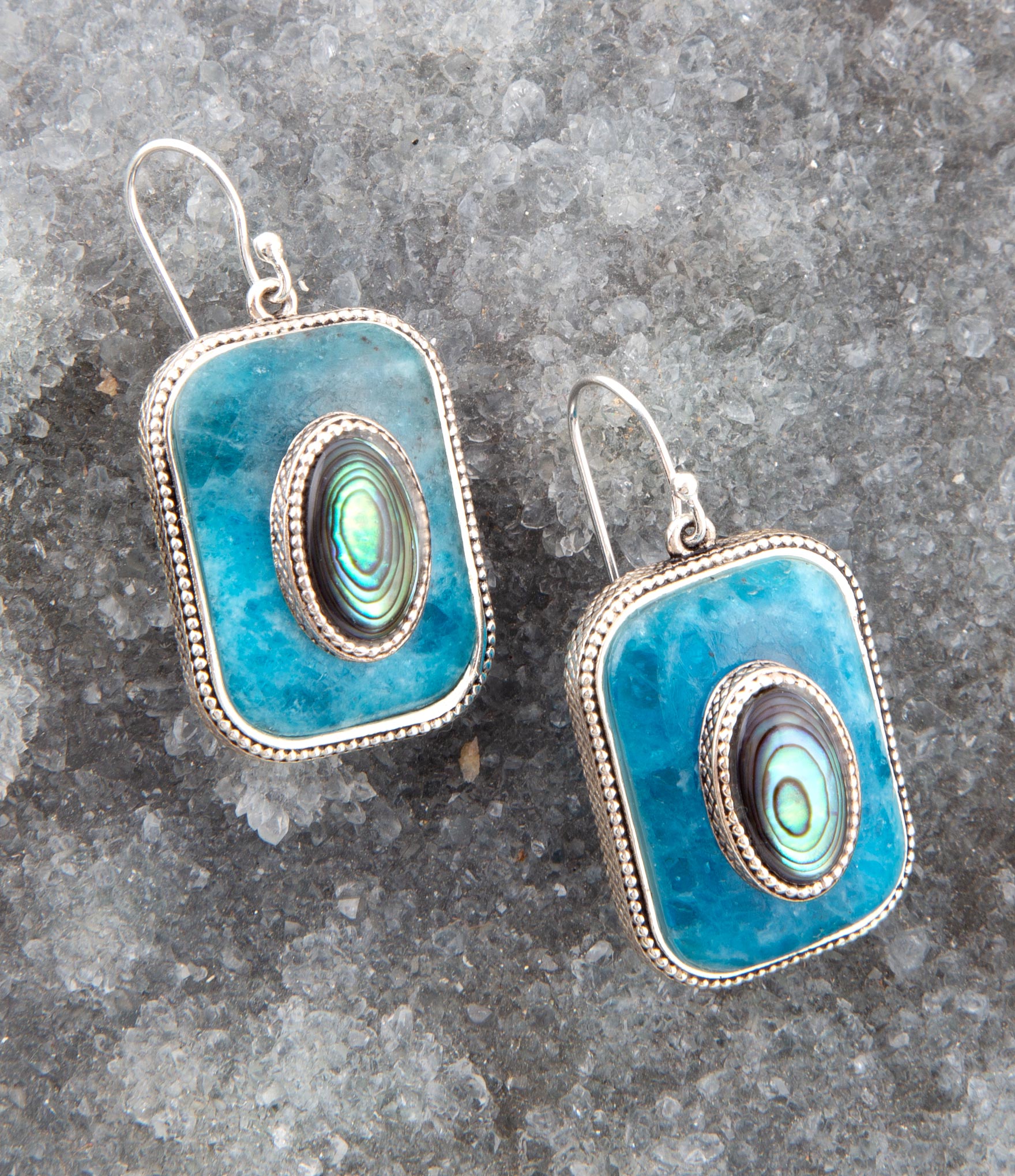 Pair of earrings with blue square design and abalone shell center on a gray stone surface
