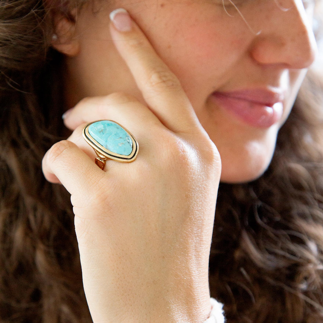 A close-up lifestyle shot of a woman wearing a gold ring with a large, rounded turquoise-blue cabochon stone on her finger.