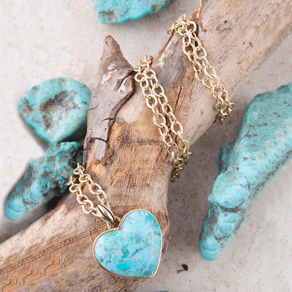 Gold necklace with a turquoise heart pendant on a wooden surface with turquoise stones.