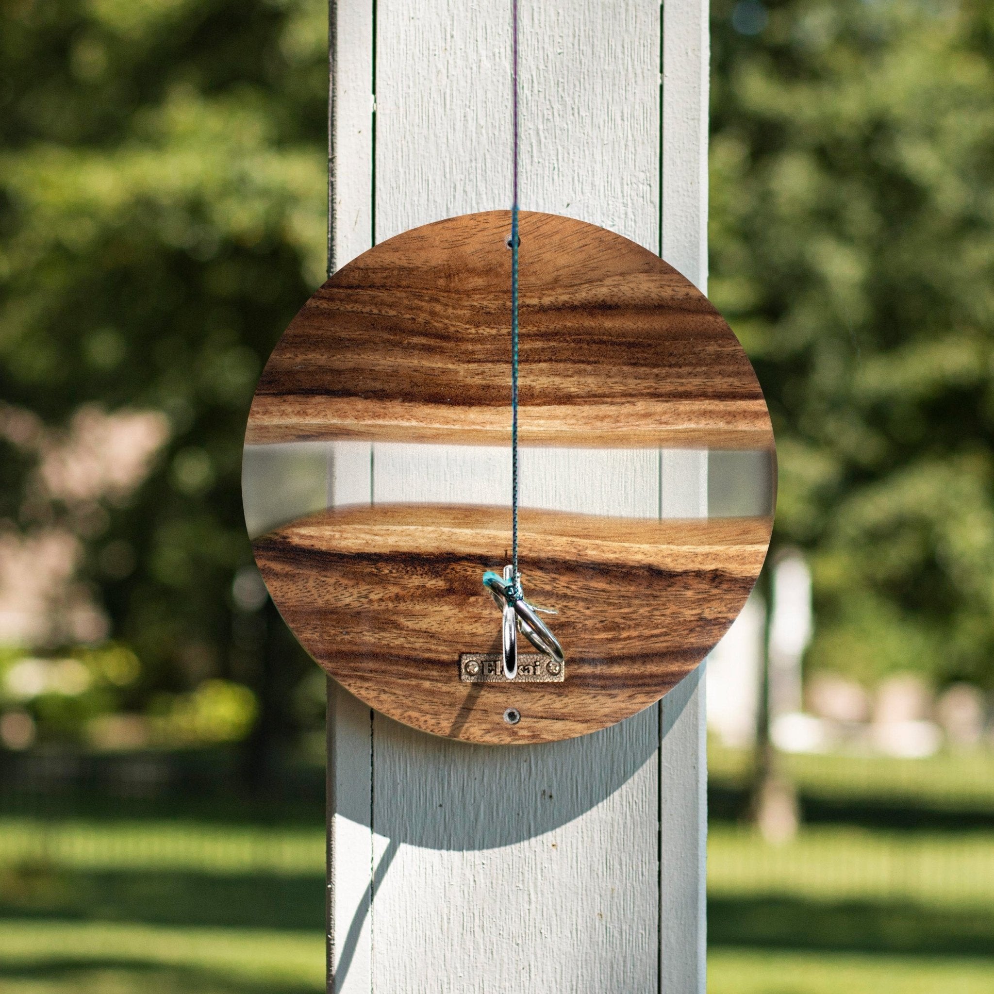 A round wooden wall clock with a reflective mirror face and a small metal bird perched on the center, hanging outdoors on a white wooden post with a blurred green garden background.