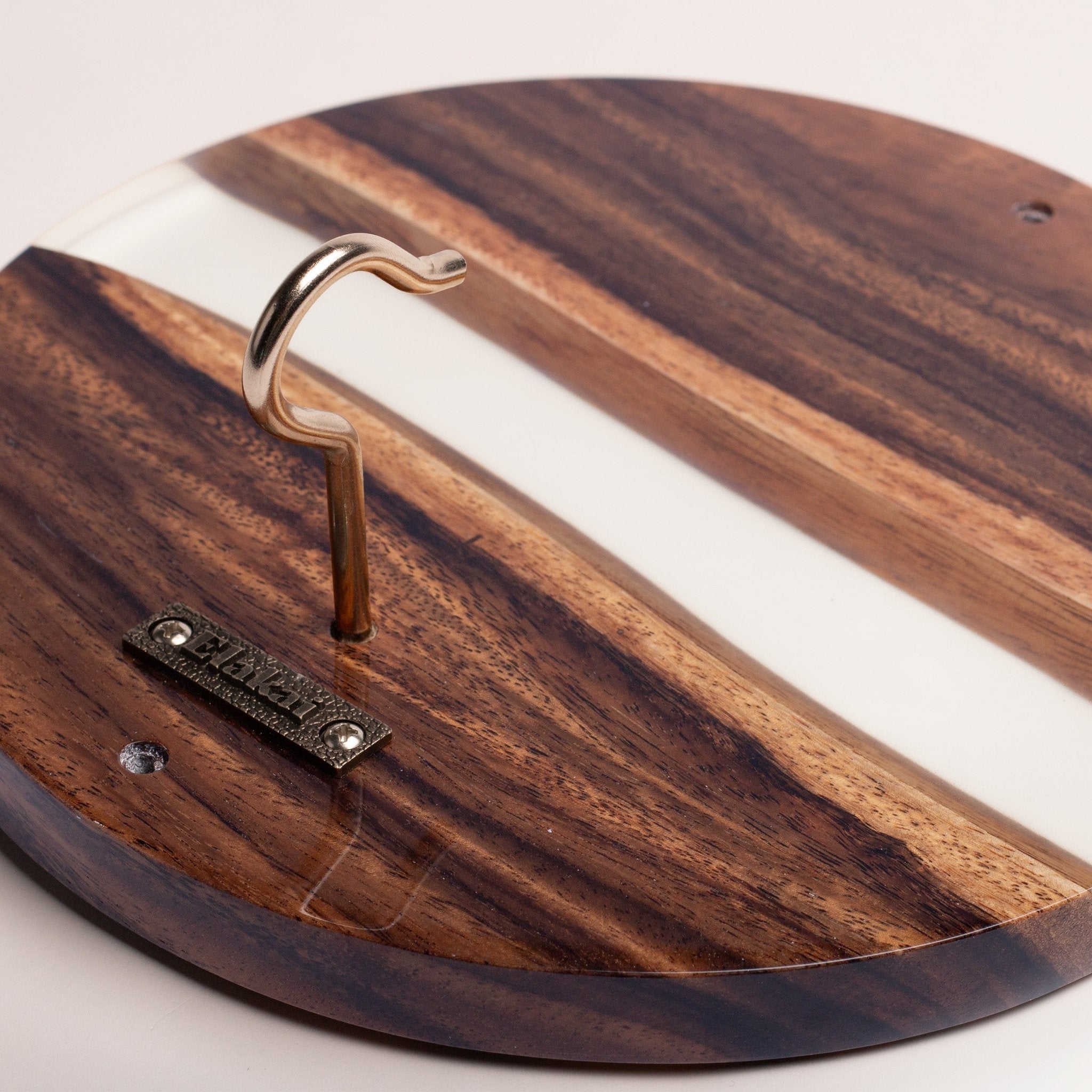 A close-up of a round wooden board with a natural dark wood grain pattern and a white inlay stripe. A curved metal hook is mounted on the board, along with a small metal brand plaque that reads 'PINECRAFT'.