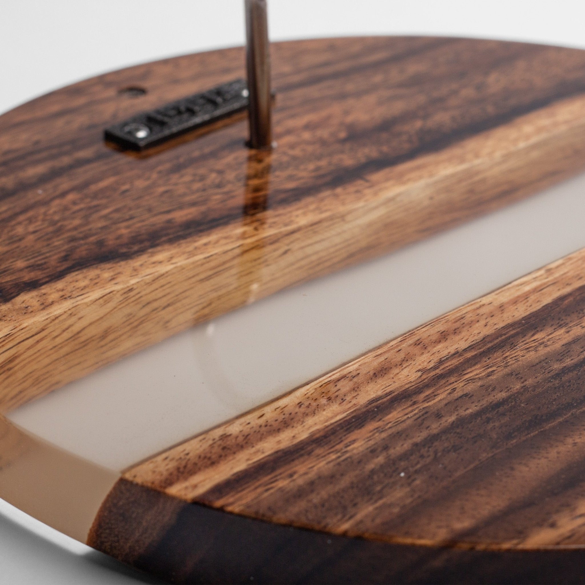 Close-up of a round wooden tray or board with a glossy dark brown wood grain finish, a translucent beige resin inlay, and a small black metal brand plaque near the top edge.